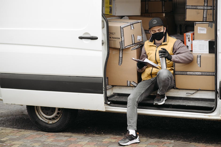 Masked courier sitting in a van, reviewing documents, surrounded by delivery boxes.